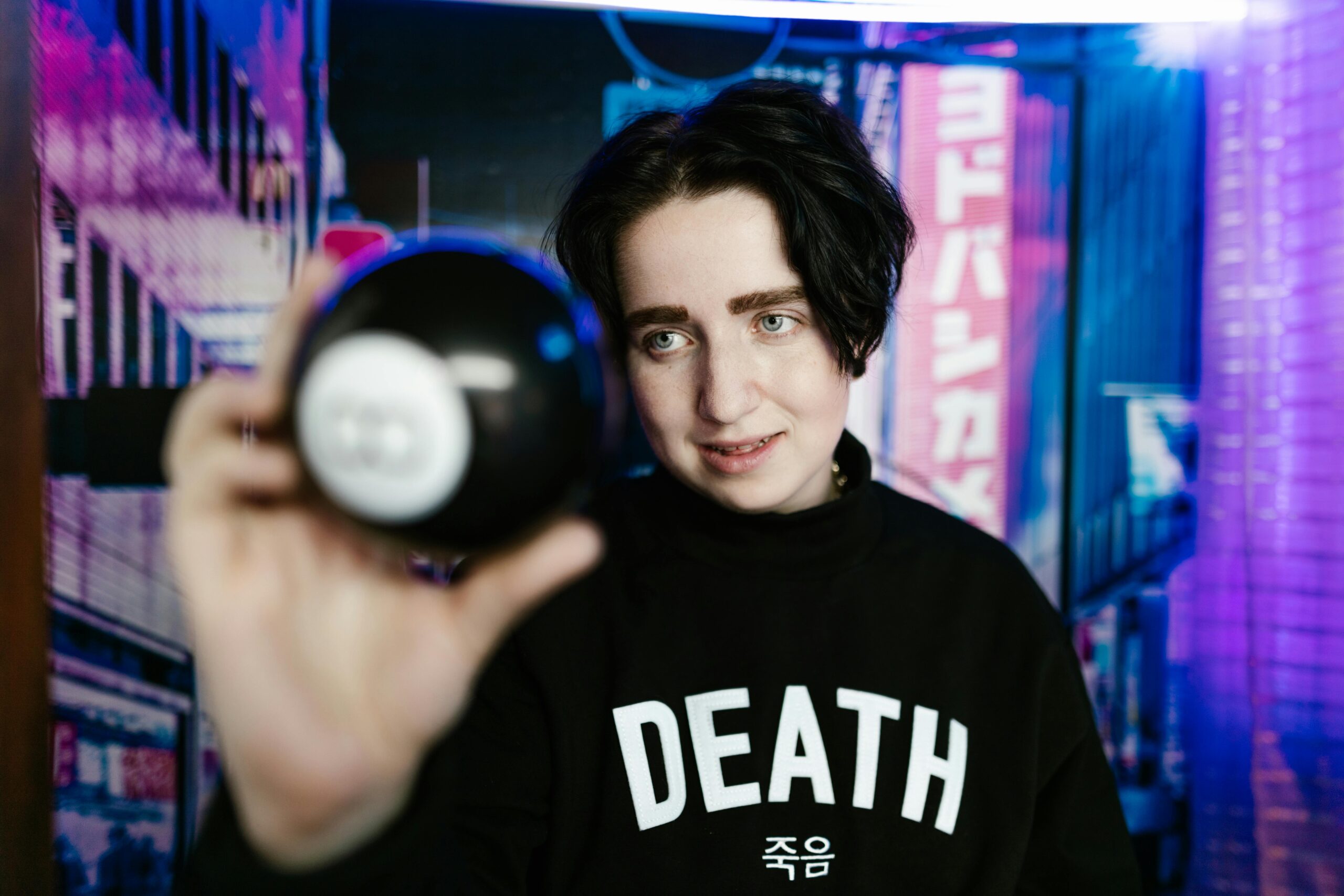 A person interacts with a magic 8 ball in a vibrant neon-lit cityscape, wearing a 'Death' sweatshirt.
